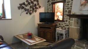A living room with a stone wall, a flat-screen TV, a fireplace, and a coffee table with a vase of flowers. - GITE 3 EPIS BETWEEN LAMBALLE AND PLANCOET FOR 8 PERSONS (saint denoual)