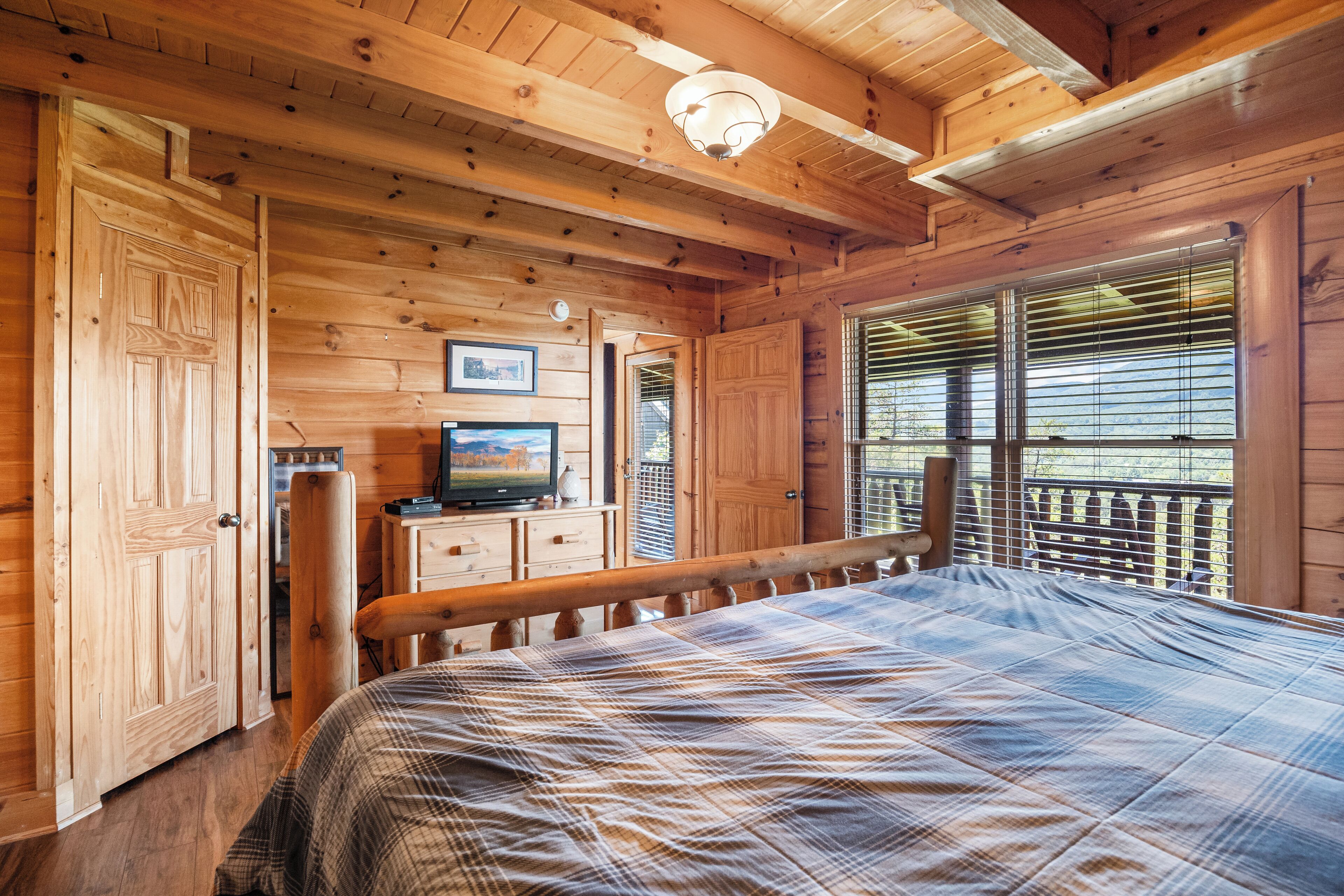 A wooden cabin room with a bed, a dresser, a television, and a window with blinds.