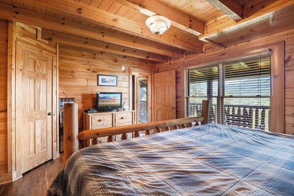 Une chambre dans un chalet en bois, comprenant un lit, une commode, un téléviseur et une fenêtre avec des stores.