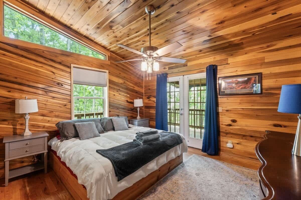 A wooden cabin bedroom with a bed, two bedside tables, a ceiling fan, and a large window.