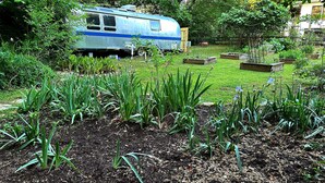 A garden with a blue RV in the background, surrounded by greenery and a stone border.