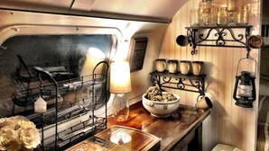 A compact kitchen with a metal rack holding dishes, a bowl on a wooden table, and a shelf with jars and a lantern.