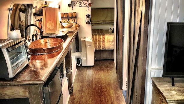 A compact kitchen with a stove, sink, and a small refrigerator. There is a bed with a striped blanket and a window with curtains.