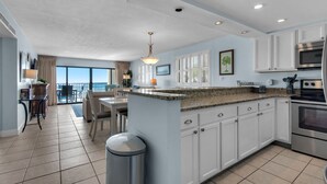 Una cocina moderna con gabinetes blancos, encimera de granito y electrodomésticos de acero inoxidable. Hay un comedor con mesa y sillas, y vistas al océano a través de amplias ventanas.