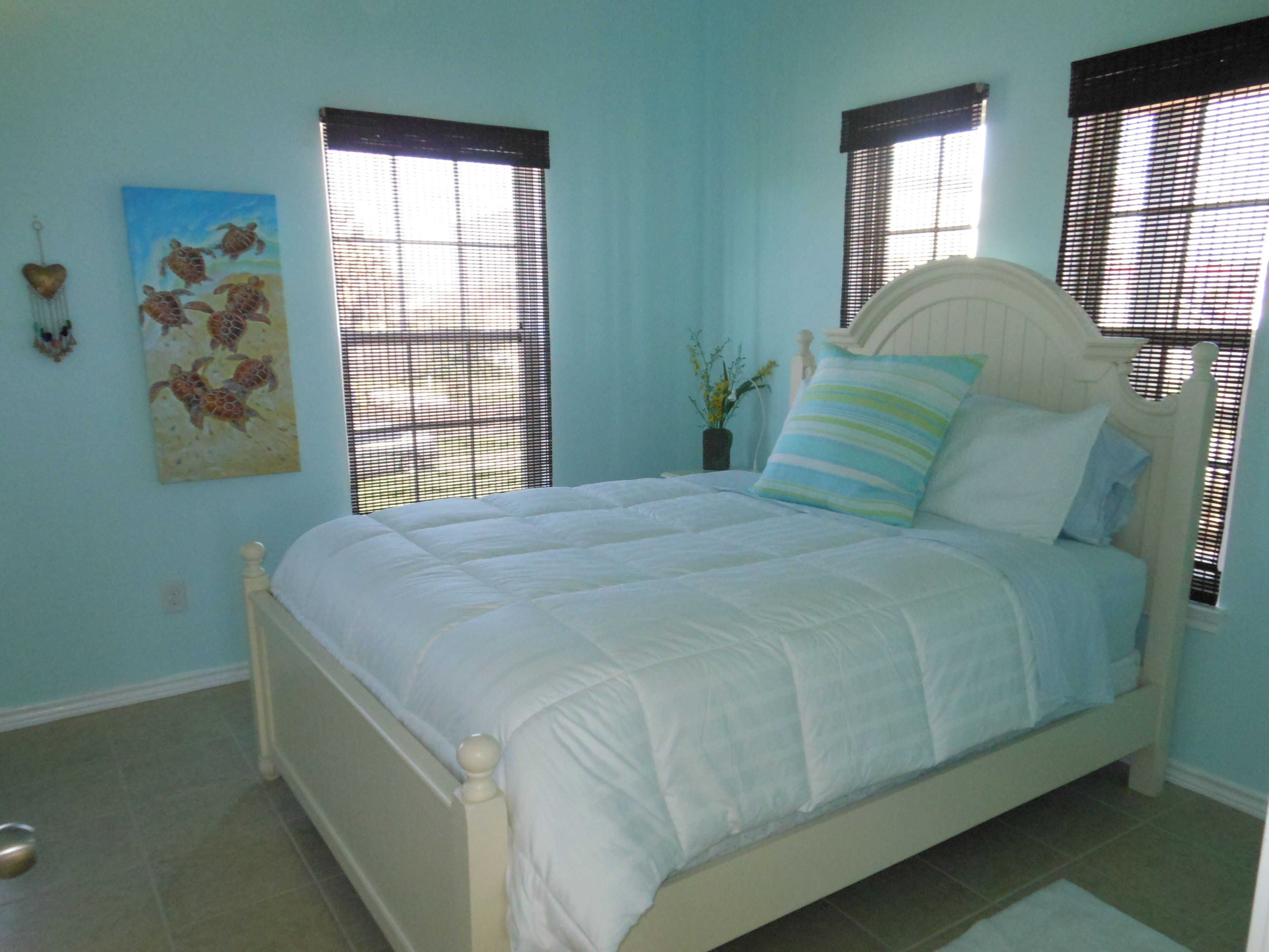 A bedroom with a bed, two windows with blinds, a wall-mounted painting, and a small plant.