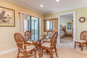 A dining area with a glass-top table surrounded by wicker chairs, a framed picture on the wall, and a view into a bedroom through an open door.