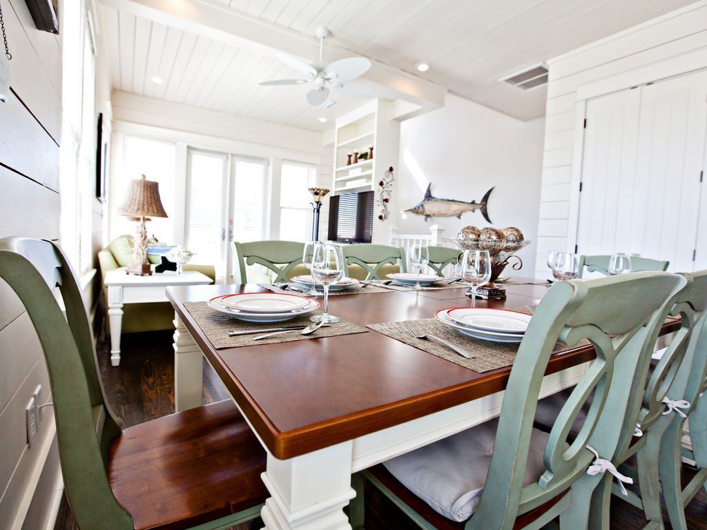 A dining area with a wooden table set for four, surrounded by green chairs. A fish mural adorns the wall, and a ceiling fan is visible.