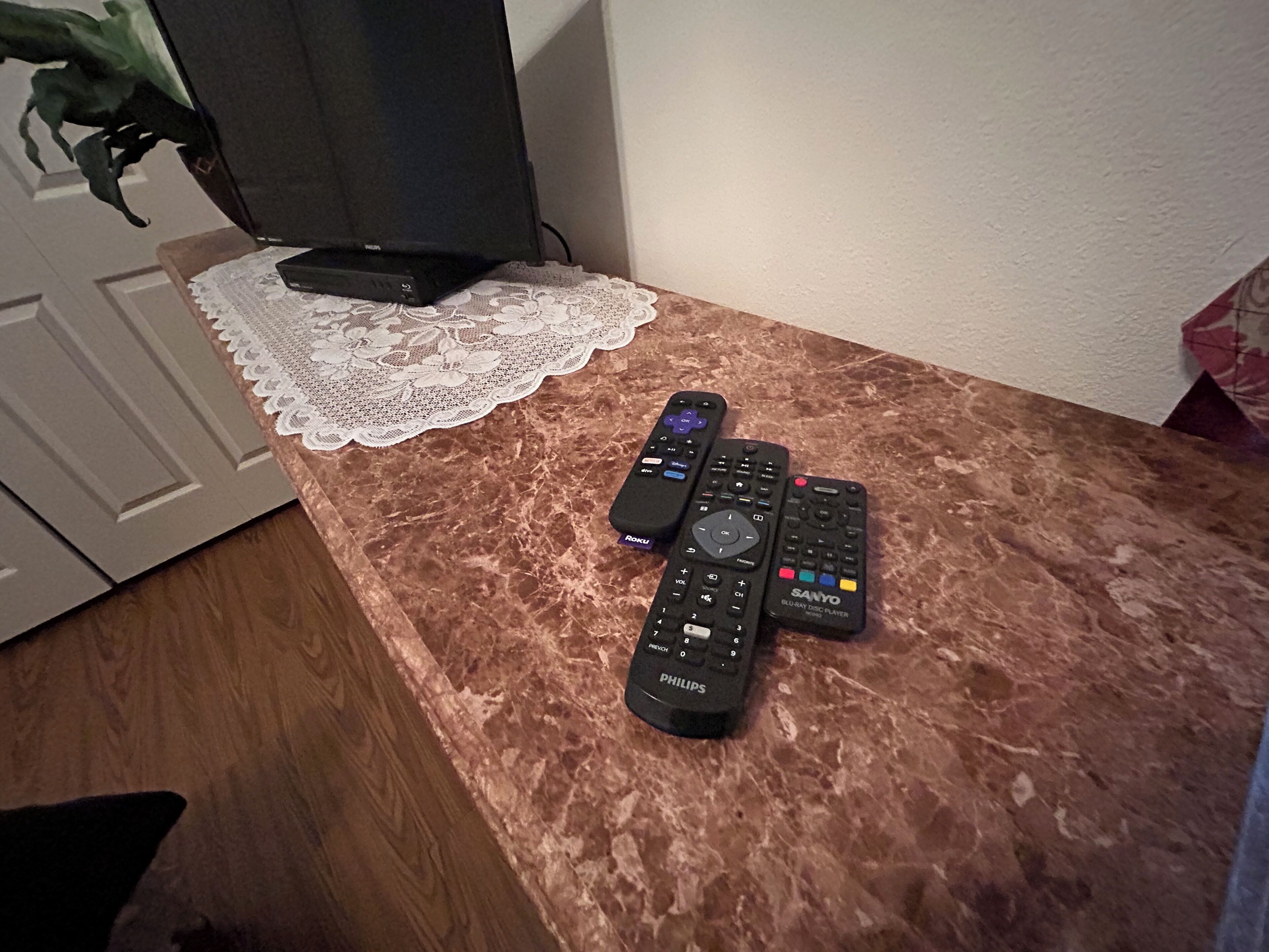Eine Marmorarbeitsplatte mit drei Fernbedienungen, einem schwarzen Fernseher und einer Spitzeneinlage.