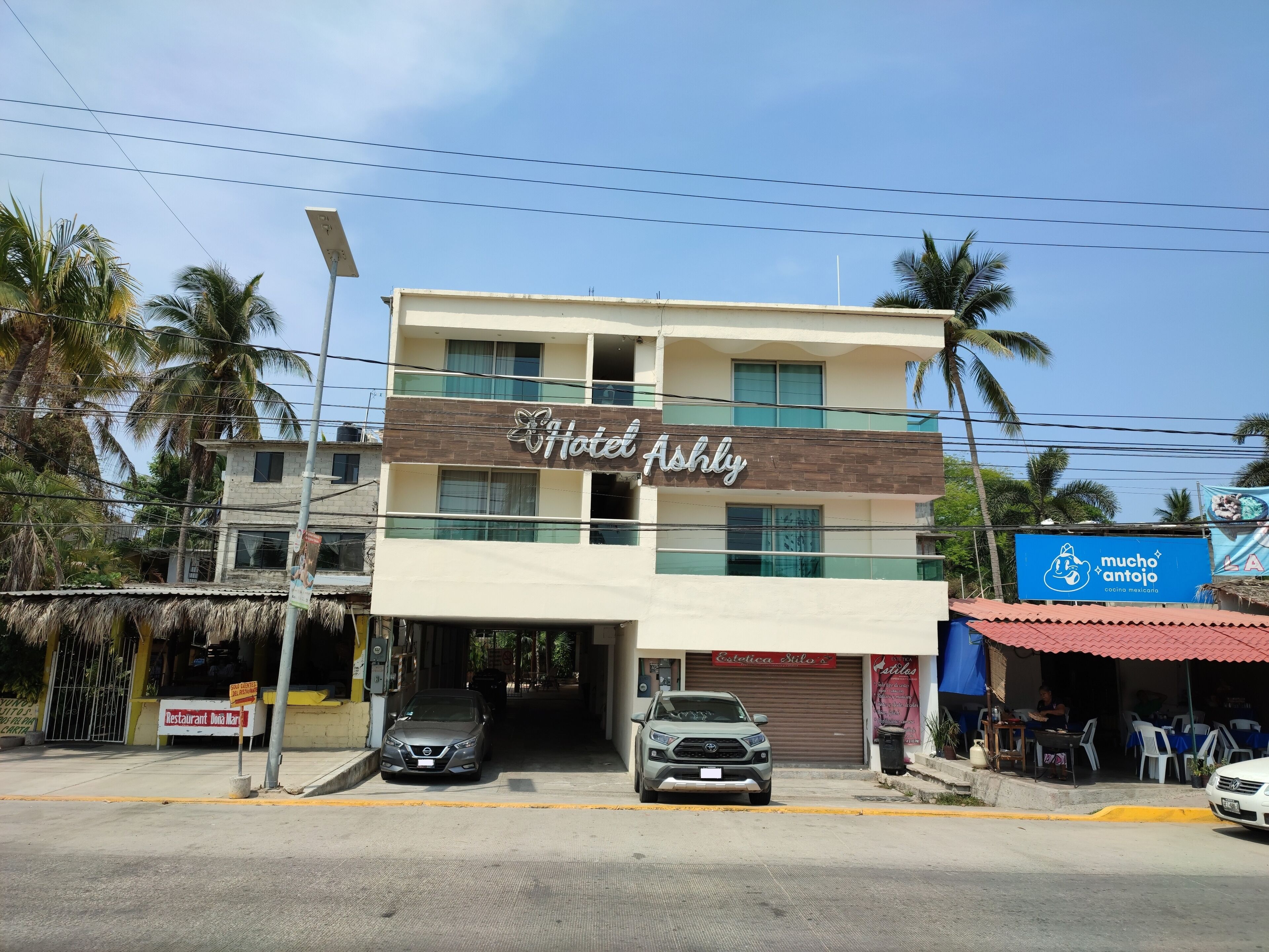Un hotel llamado "Hotel Ashly" con un diseño moderno, una entrada cubierta y un restaurante visible llamado "Dona Mart" al fondo.