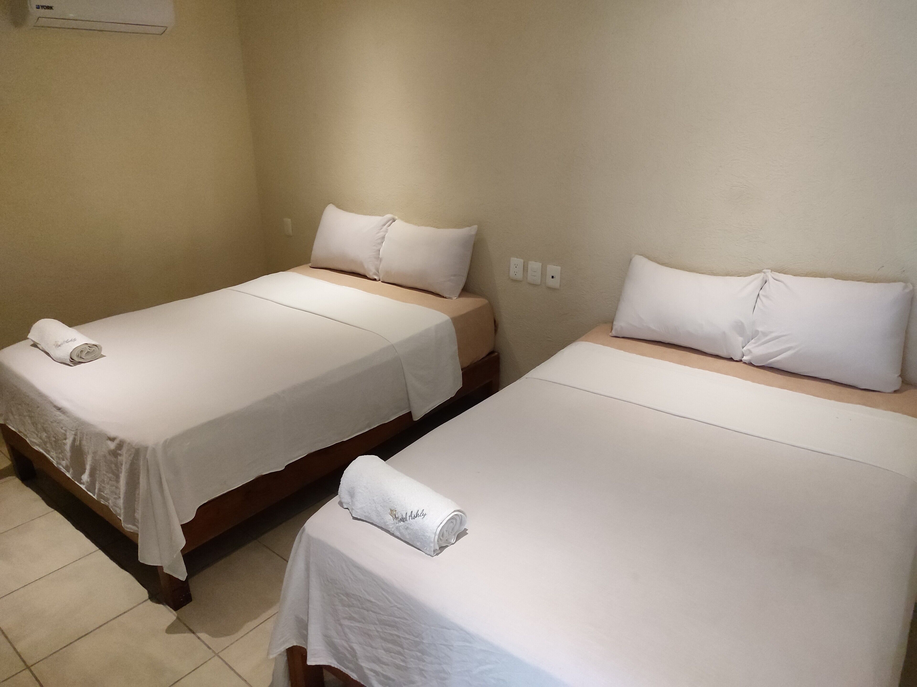 Dos camas individuales con sábanas y almohadas blancas, un cabecero de madera y una mesita con una toalla.