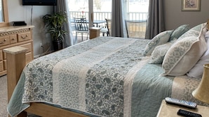 A bedroom with a large bed, a television on the wall, a desk with a lamp, and a balcony with a table and chairs.