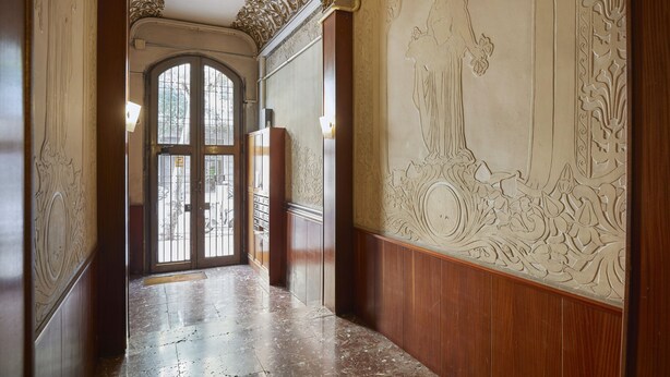 A hallway with ornate wall carvings, a wooden floor, and a large arched doorway leading outside.