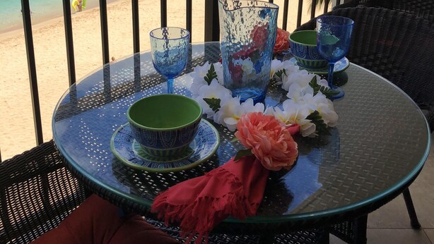 A round glass table with a green bowl, blue glassware, and a red cloth on a balcony overlooking a beach. - Stay On The Beach In Makaha   (Waianae)