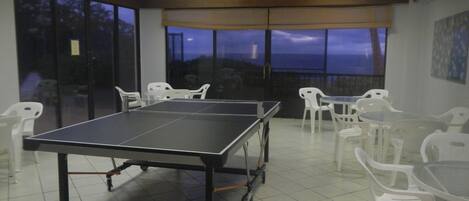 Uma mesa de pingue-pongue em um espaço com cadeiras brancas, grandes janelas e vista do céu.
