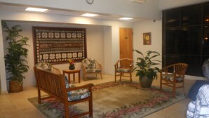A room with a wooden sofa, two armchairs, a large decorative wall hanging, a potted plant, and a door. - Stay On The Beach In Makaha   (Waianae)
