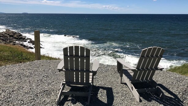 Two Adirondack chairs facing the ocean with a rocky shoreline and a wooden post in the background.