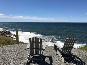 Deux fauteuils Adirondack sont placés face à l’océan, avec une côte rocheuse et un poteau en bois en arrière-plan.