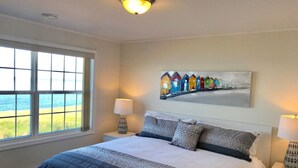 A bedroom with a large bed, two bedside tables with lamps, a window with a view of the beach, and a painting of colorful beach houses on the wall.