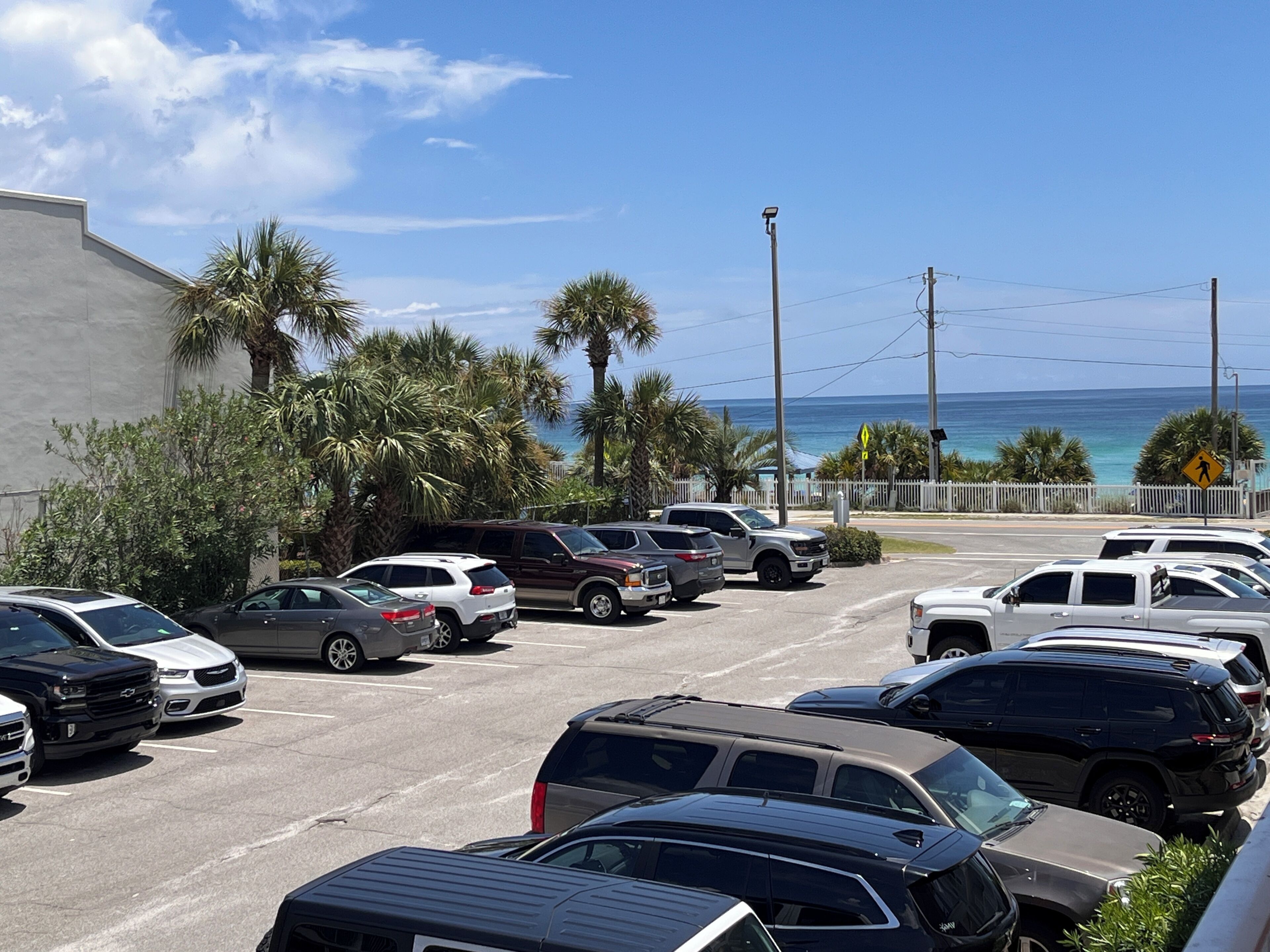A parking lot with multiple cars, a white building, palm trees, and a view of the ocean.