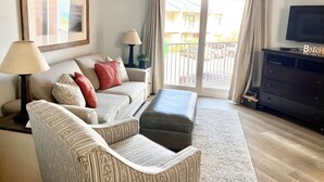 A living room with a sofa, ottoman, lamp, TV, and a ceiling fan. - Private Beach & Free Beach Service; Walkable Scenic 98 to Dining; Pool & Hot Tub (Miramar Beach)