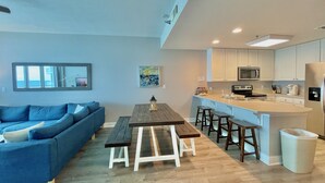 Een moderne woonkamer met een blauwe bank, een houten eettafel met bankjes, een keuken met witte kastjes en roestvrijstalen apparatuur, en een groot raam met uitzicht.