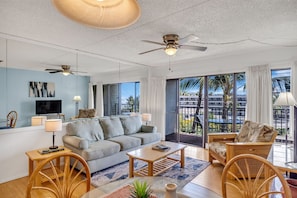 A spacious living room with a sofa, coffee table, and chairs, large windows with a view of palm trees, ceiling fans, and a flat-screen TV.