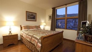 A bedroom with a wooden bed, two bedside tables with lamps, a window with a view of buildings, and a framed picture on the wall. - Granita 301 (Telluride)