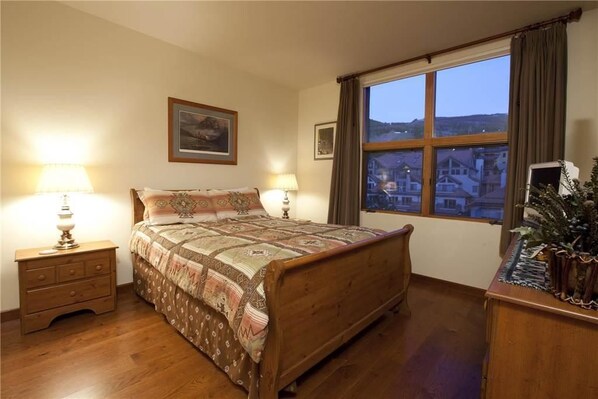 A bedroom with a wooden bed, two bedside tables with lamps, a window with a view of buildings, and a framed picture on the wall. - Granita 301 (Telluride)