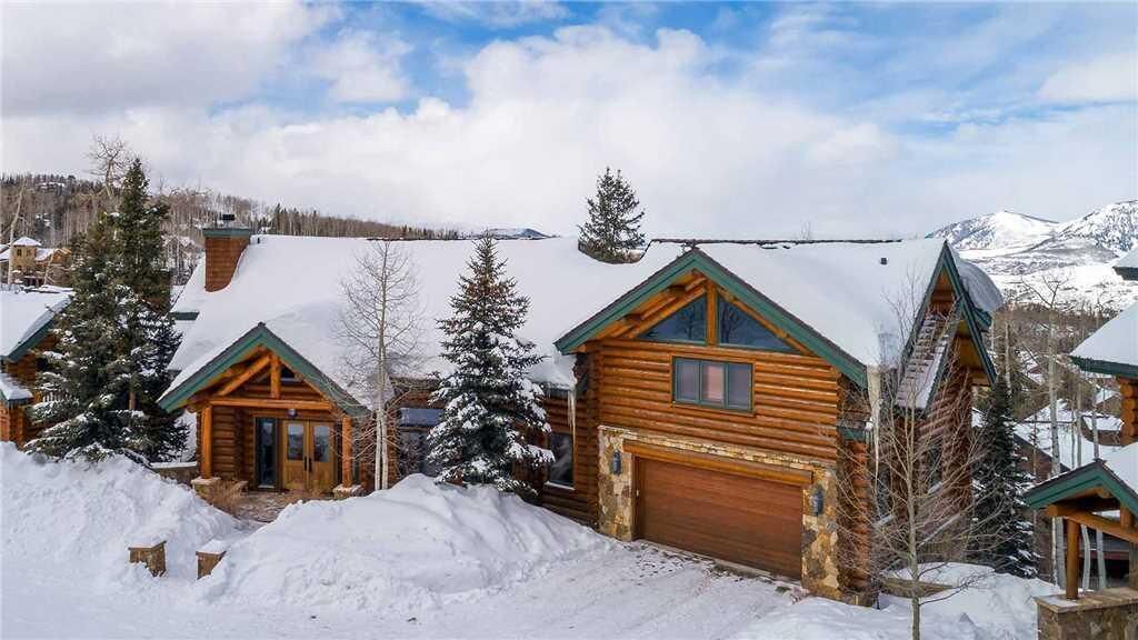 A log cabin with a garage, surrounded by snow-covered trees and mountains in the background.