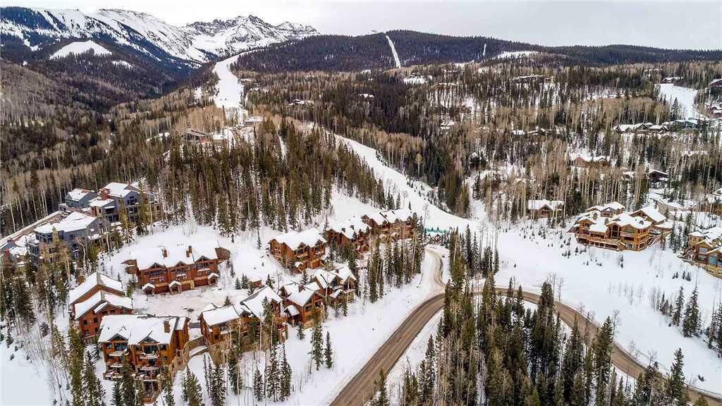 A snow-covered lodge with multiple buildings, a winding road, and a mountainous backdrop.