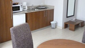 Eine moderne Küche mit Mikrowelle, Kaffeemaschine und einer kleinen Arbeitsfläche. Es gibt einen Holzschrank, einen Mülleimer und einen kleinen Tisch mit einer Zeitschrift.