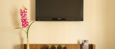 A flat-screen TV mounted on a wall above a wooden console table with a vase of flowers, a remote control, and a small photo frame.