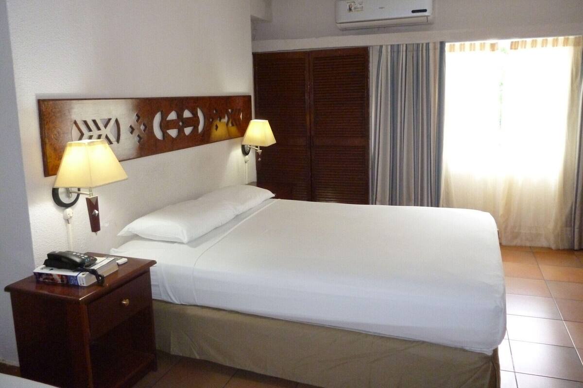 Une chambre d’hôtel comprenant un lit, deux lampes de chevet, une tête de lit en bois, une armoire, un bureau avec un téléphone et un appareil de climatisation.