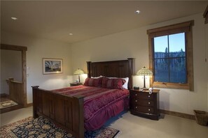 A bedroom with a wooden bed, two bedside lamps, a nightstand, a window with a view of trees, and a framed picture on the wall.