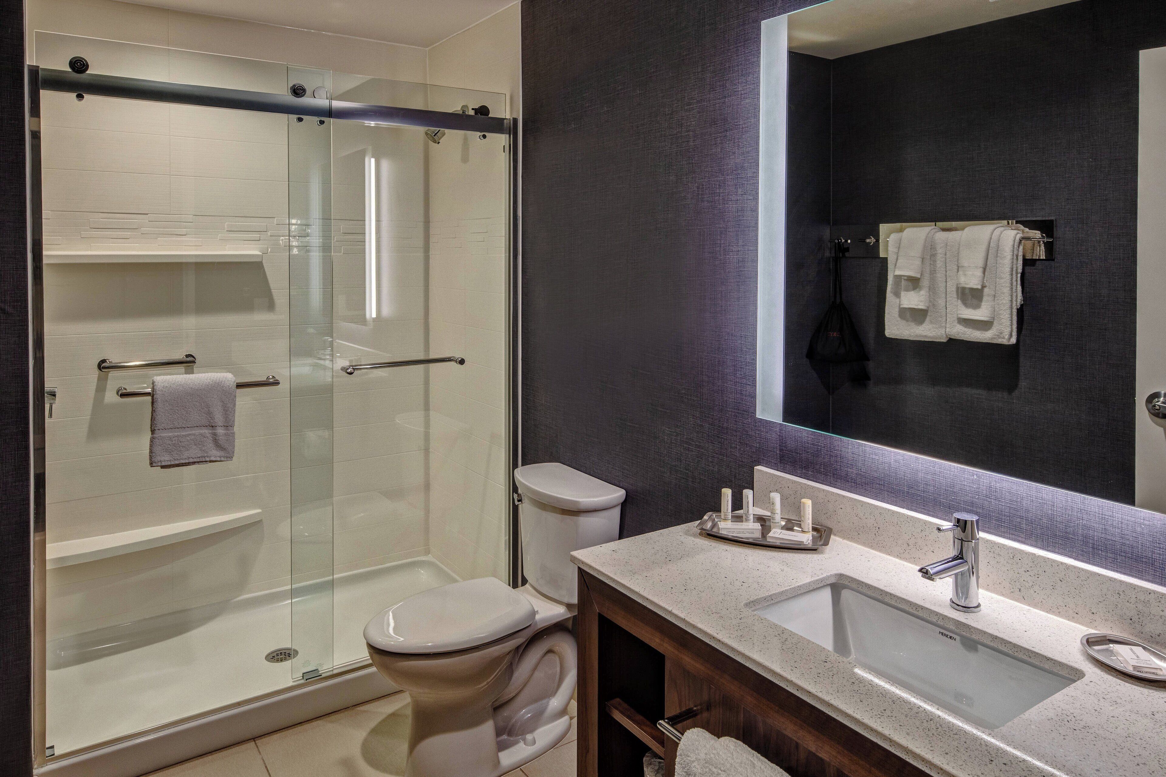 A modern bathroom with a glass-enclosed shower, a white toilet, a large mirror, and a countertop with toiletries.