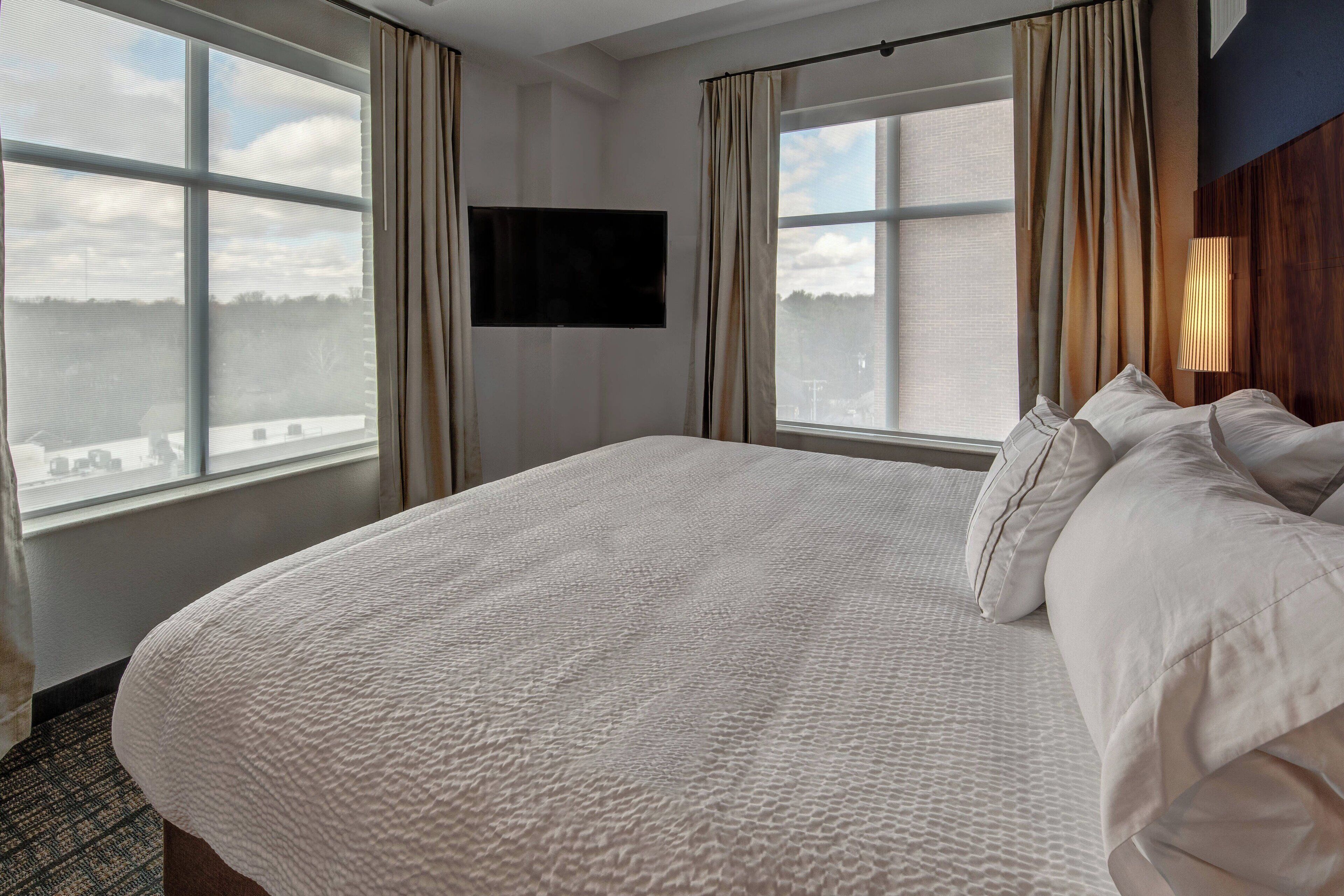 A hotel room with a large bed, two windows with curtains, a mounted TV, and a city view.