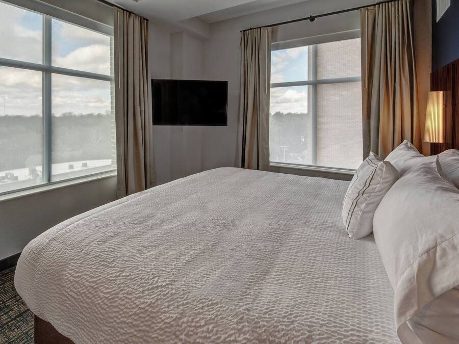 A hotel room with a large bed, two windows with curtains, a mounted TV, and a city view.