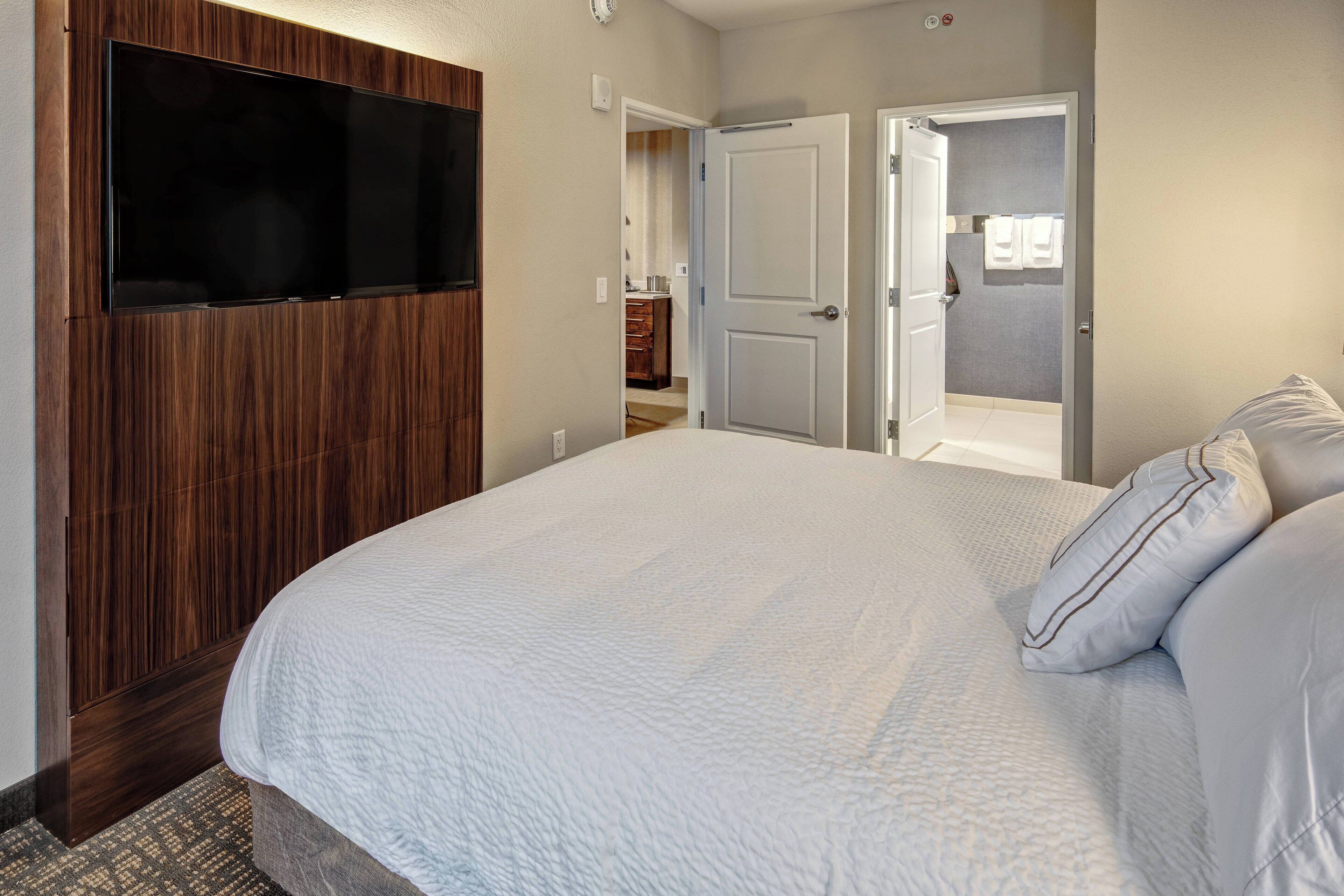A bedroom with a large bed, a TV mounted on a wooden panel, and two doors leading to other rooms.