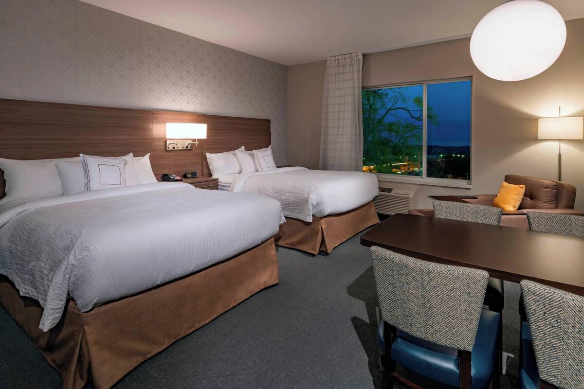 A hotel room with two beds, a dining table, and a view of the city at dusk.