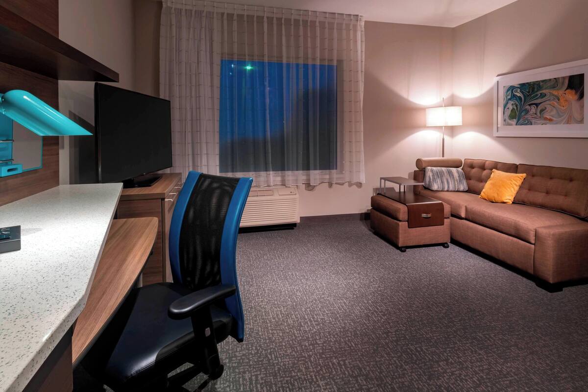 A hotel room with a desk, a blue office chair, a brown sofa with a yellow pillow, a standing lamp, and a large window with curtains.
