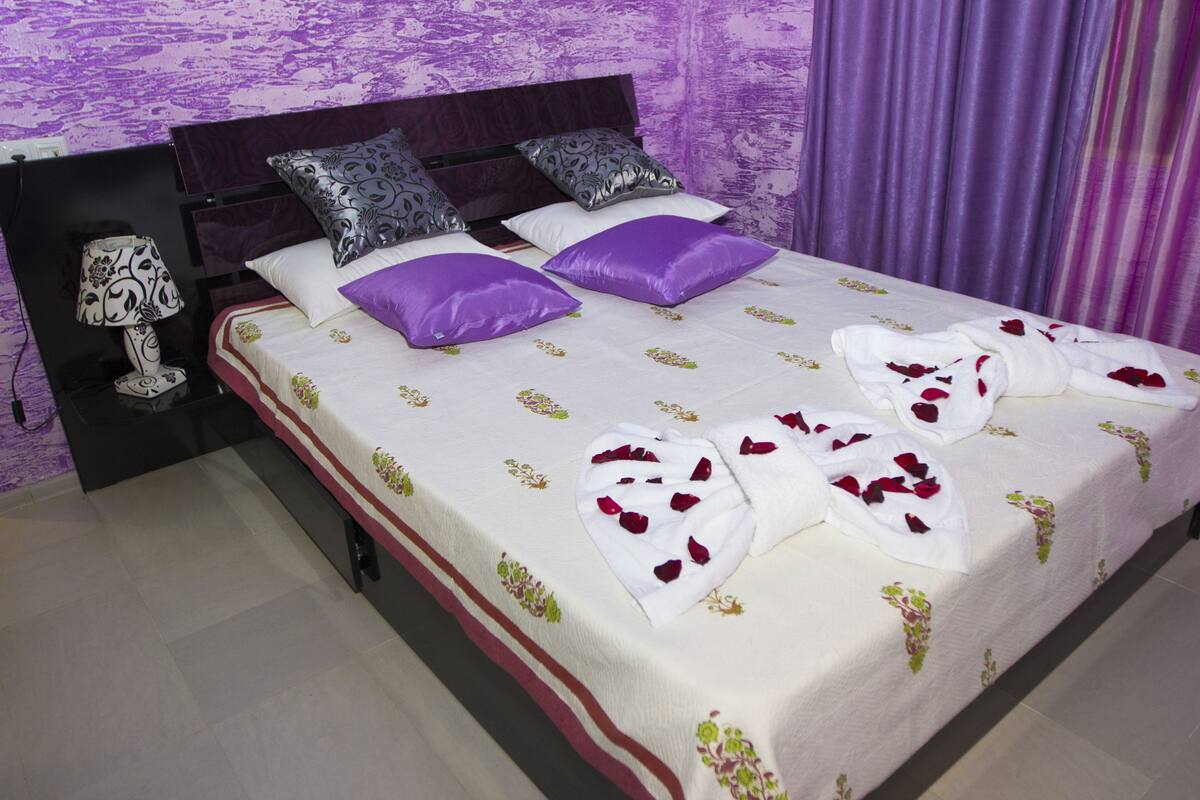 Un lit bien fait, recouvert d’une couvre-lit à motifs floraux, avec deux oreillers violets et des pétales de rose disposés sur le lit.