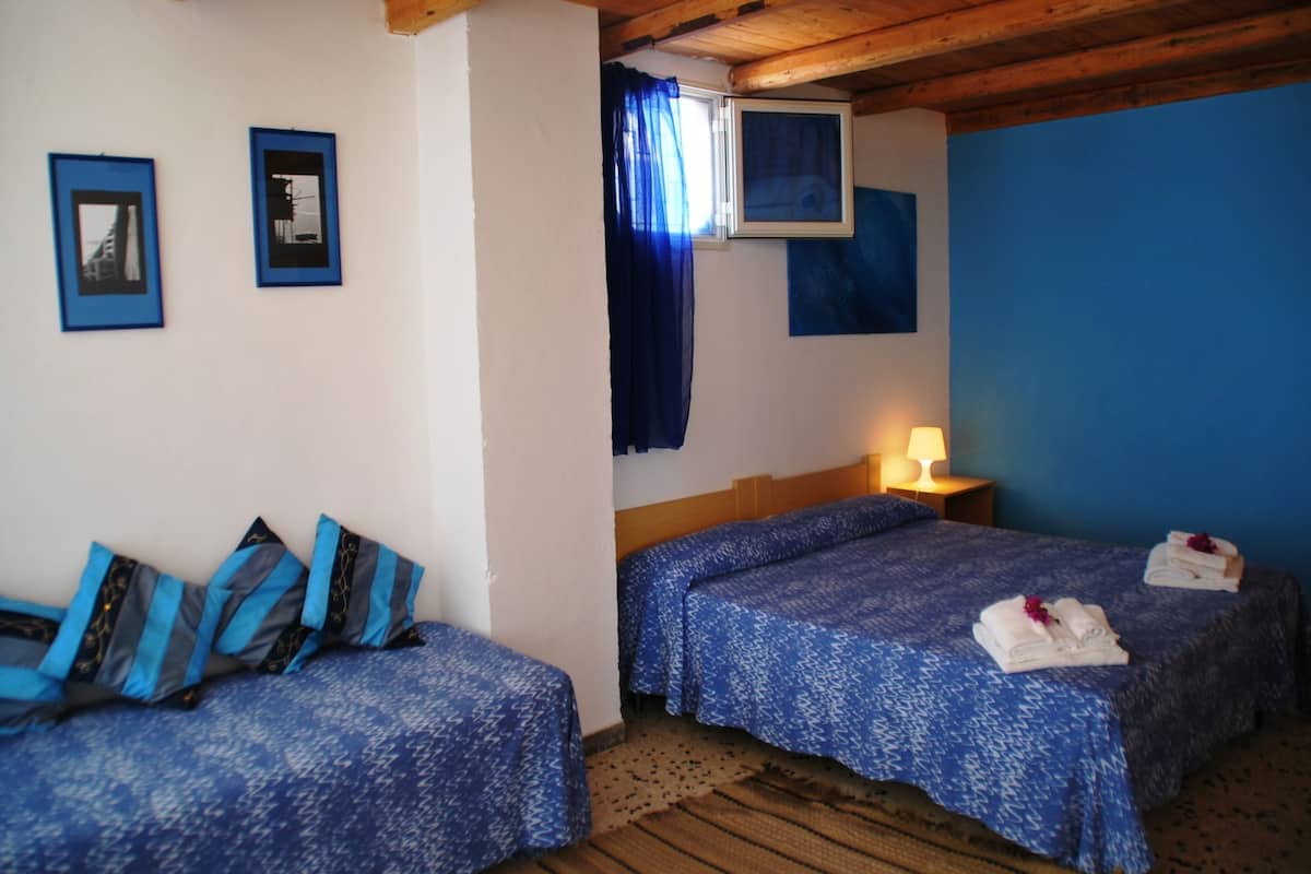 A room with two beds, a window with blue curtains, a wall-mounted TV, and a lamp on a bedside table.