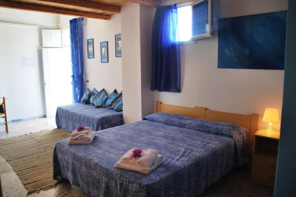 A hotel room with two beds, a wooden headboard, a nightstand with a lamp, and a window with blue curtains.