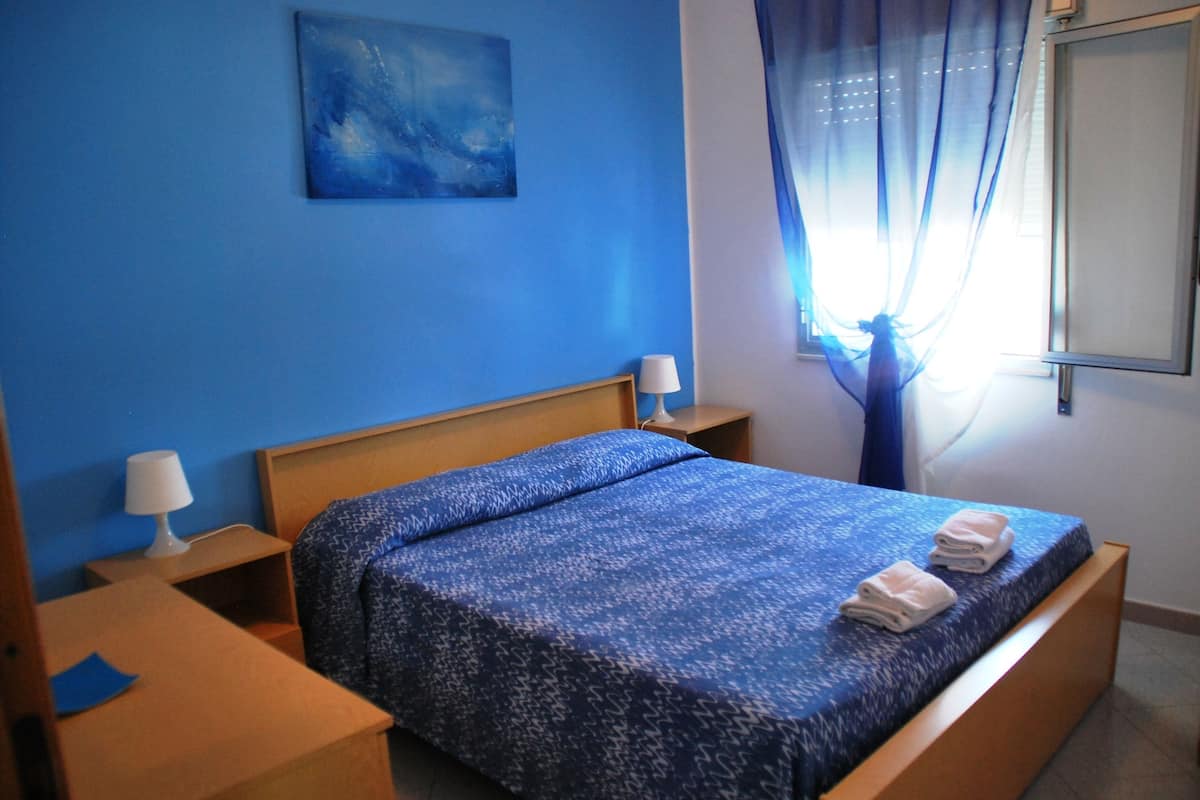 A hotel room with a single bed, a nightstand, a lamp, a painting on the wall, and a window with curtains.