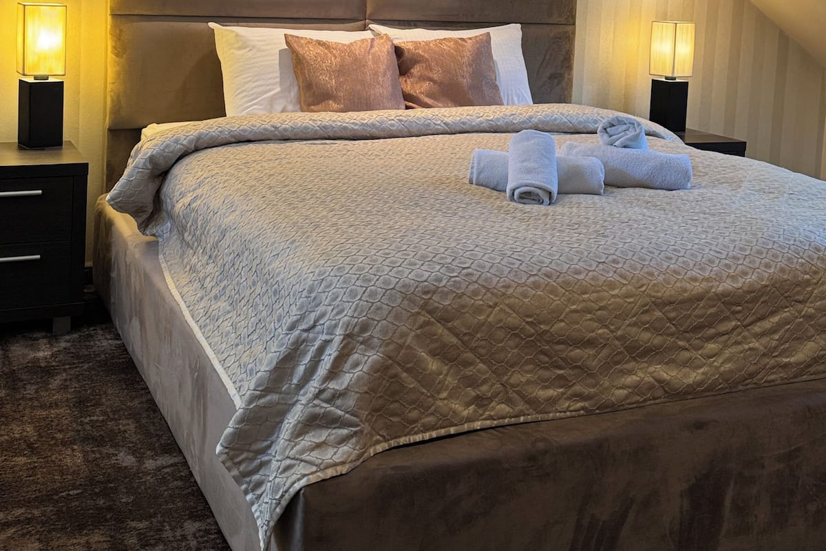 A bedroom with a sloped ceiling, a large bed with a quilted cover, two bedside lamps, and a nightstand.
