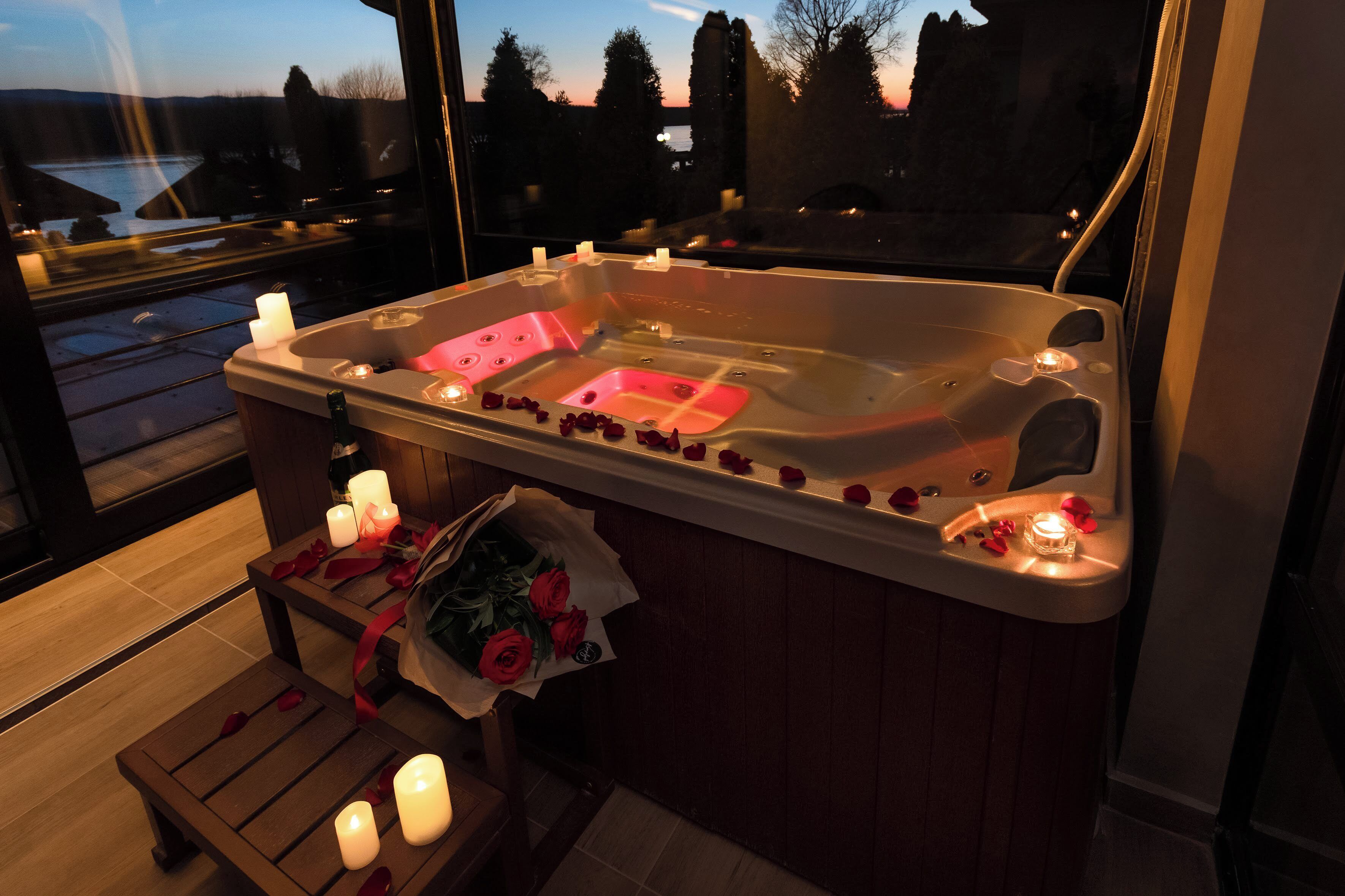 Una vasca idromassaggio con petali di rosa e candele, una panca in legno e vista su alberi e uno specchio d'acqua.