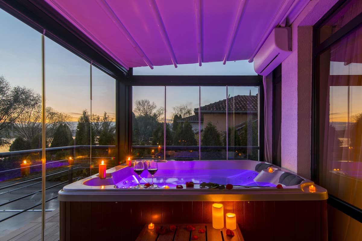 A hot tub with candles and wine glasses on a balcony with a view of trees and houses.