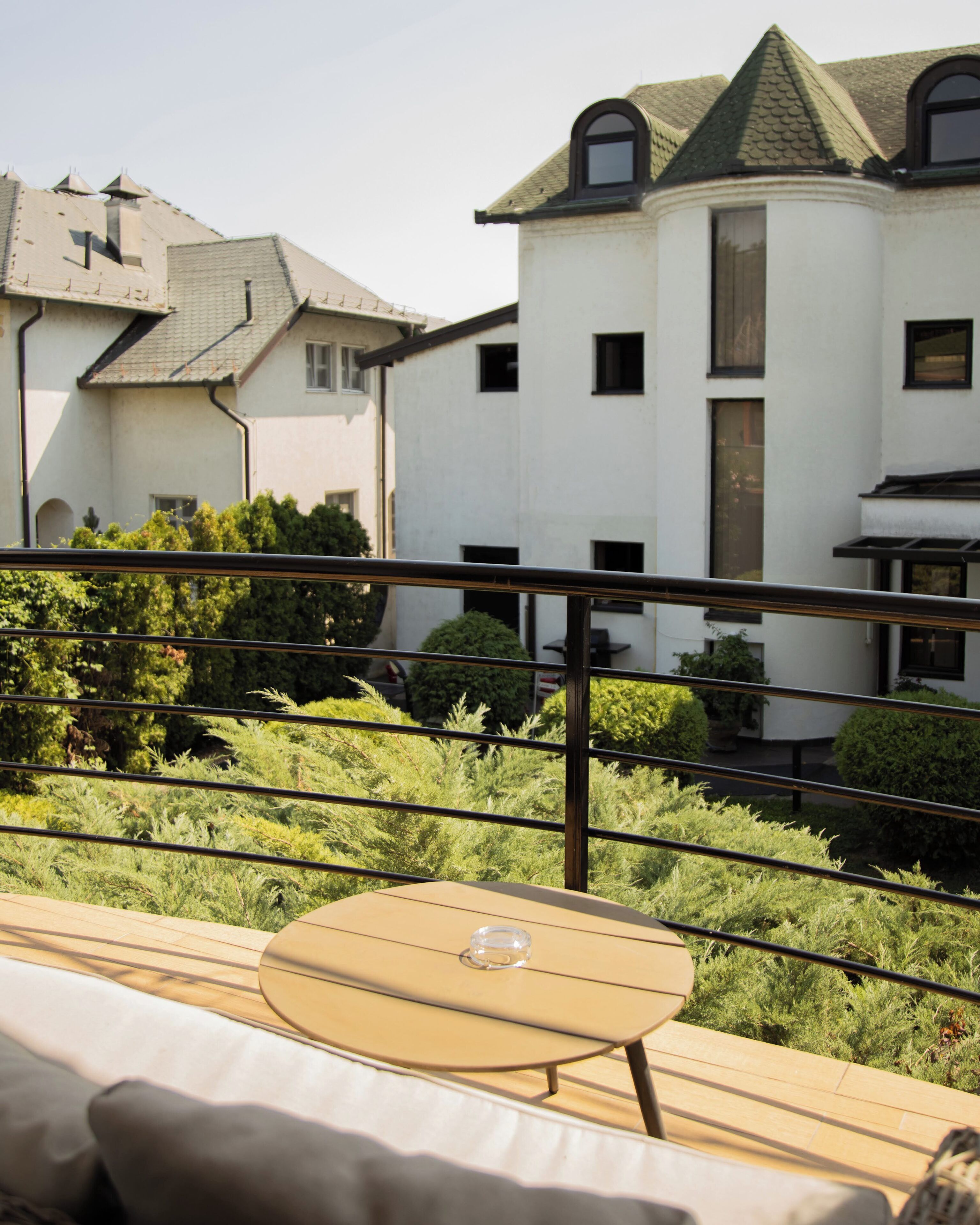A balcony with a table and chairs, surrounded by greenery and overlooking a building with multiple windows and a distinctive roof.