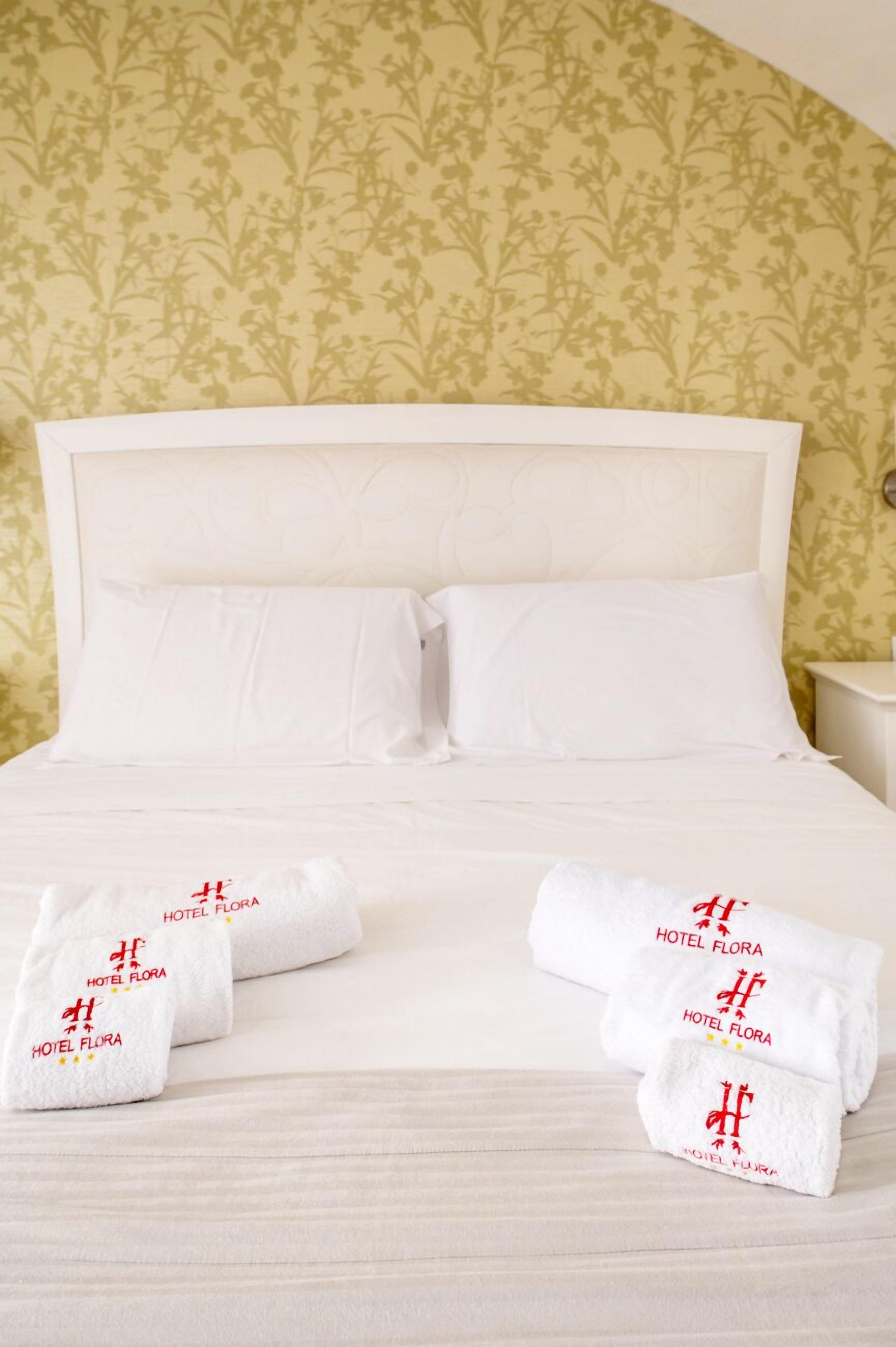 A neatly made bed with white linens and two towels with 'HOTEL FLORA' branding.