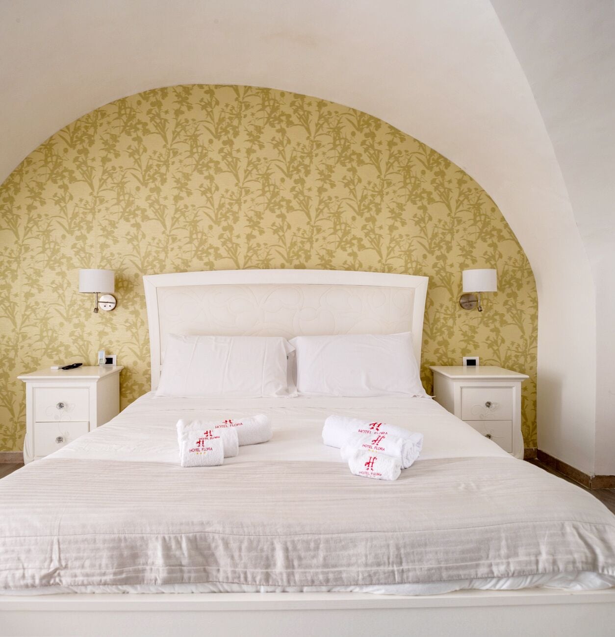 Una cama bien tendida con sábanas y almohadas blancas, dos mesitas de noche con lámparas, y un papel tapiz con motivos florales.